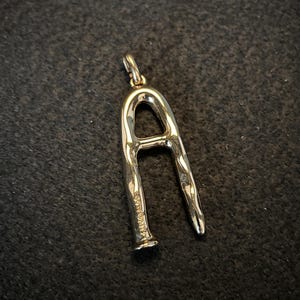 Puede incluir: Un colgante plateado con forma de la letra "A". El colgante tiene una superficie texturizada y un peque&ntilde;o lazo en la parte superior para una cadena. La letra est&aacute; estilizada con una forma ligeramente irregular.