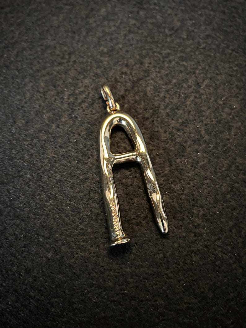 Puede incluir: Un colgante plateado con forma de la letra "A". El colgante tiene una superficie texturizada y un peque&ntilde;o lazo en la parte superior para una cadena. La letra est&aacute; estilizada con una forma ligeramente irregular.