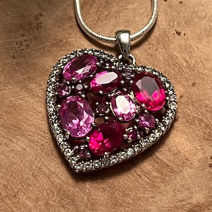 May include: A heart-shaped pendant necklace featuring a cluster of oval and round faceted pink and deep red gemstones set in silver. The heart frame is bordered by a row of small, clear, sparkling stones, and the pendant hangs from a silver chain.