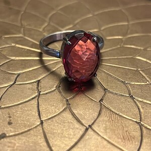 Pode incluir: Um anel de prata com uma grande pedra preciosa vermelha oval e facetada. A pedra preciosa &eacute; o ponto focal, captando a luz e exibindo uma cor vibrante. A banda do anel &eacute; simples, permitindo que a pedra preciosa se destaque.