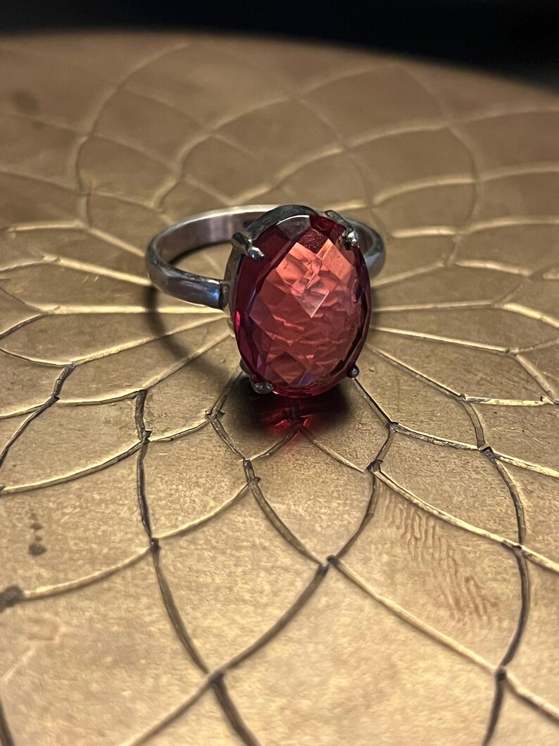 Pode incluir: Um anel de prata com uma grande pedra preciosa vermelha oval e facetada. A pedra preciosa &eacute; o ponto focal, captando a luz e exibindo uma cor vibrante. A banda do anel &eacute; simples, permitindo que a pedra preciosa se destaque.