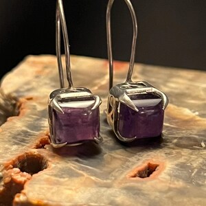 May include: A pair of silver-toned earrings featuring square, deep purple gemstones. The earrings have a hook design for pierced ears. The gemstones are set in a claw-like setting. The earrings are displayed on a brown and tan surface.