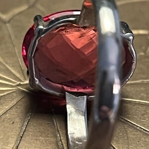 Pode incluir: Close-up de um anel de prata com uma grande pedra preciosa facetada, oval, rosa-avermelhada. A banda do anel &eacute; espessa e polida, com um design simples. A pedra preciosa &eacute; engastada numa moldura de prata.