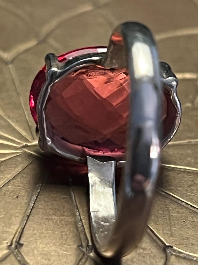 Pode incluir: Close-up de um anel de prata com uma grande pedra preciosa facetada, oval, rosa-avermelhada. A banda do anel &eacute; espessa e polida, com um design simples. A pedra preciosa &eacute; engastada numa moldura de prata.