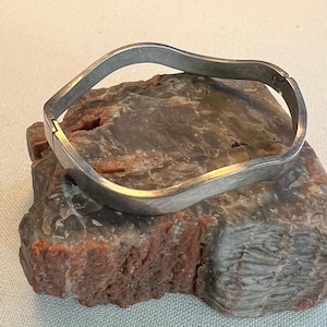 May include: A silver-colored, wavy bangle bracelet. The bracelet is hinged and appears to be made of metal. The bracelet is resting on a multi-colored, rough-textured stone.