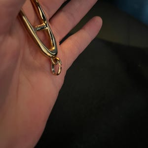 Puede incluir: Una pulsera de metal dorado con un dise&ntilde;o abierto &uacute;nico. La pulsera presenta una gran forma estilizada de la letra "A", conectada a una peque&ntilde;a cadena y un cierre. La joya se muestra sobre un fondo oscuro.