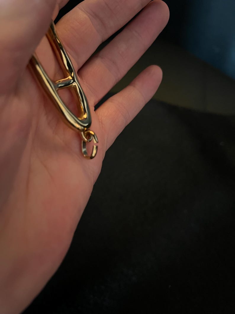 Puede incluir: Una pulsera de metal dorado con un dise&ntilde;o abierto &uacute;nico. La pulsera presenta una gran forma estilizada de la letra "A", conectada a una peque&ntilde;a cadena y un cierre. La joya se muestra sobre un fondo oscuro.
