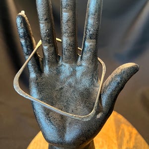 Peut inclure: Un bracelet jonc en argent de forme carrée avec une finition martelée et texturée. La bande métallique présente des coins arrondis et une surface réfléchissante, mettant en valeur un design géométrique simple d'une épaisseur constante.