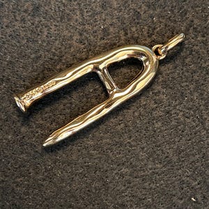 Puede incluir: Un colgante dorado con un dise&ntilde;o abstracto &uacute;nico. El colgante tiene una forma curva y abierta con una superficie texturizada y un peque&ntilde;o lazo para una cadena. El metal tiene un acabado pulido, reflejando la luz.