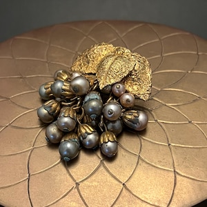 Può includere: Spilla vintage con un gruppo di perle grigie e dettagli dorati. La spilla presenta foglie dorate strutturate. Il gioiello è esposto su una superficie dorata.