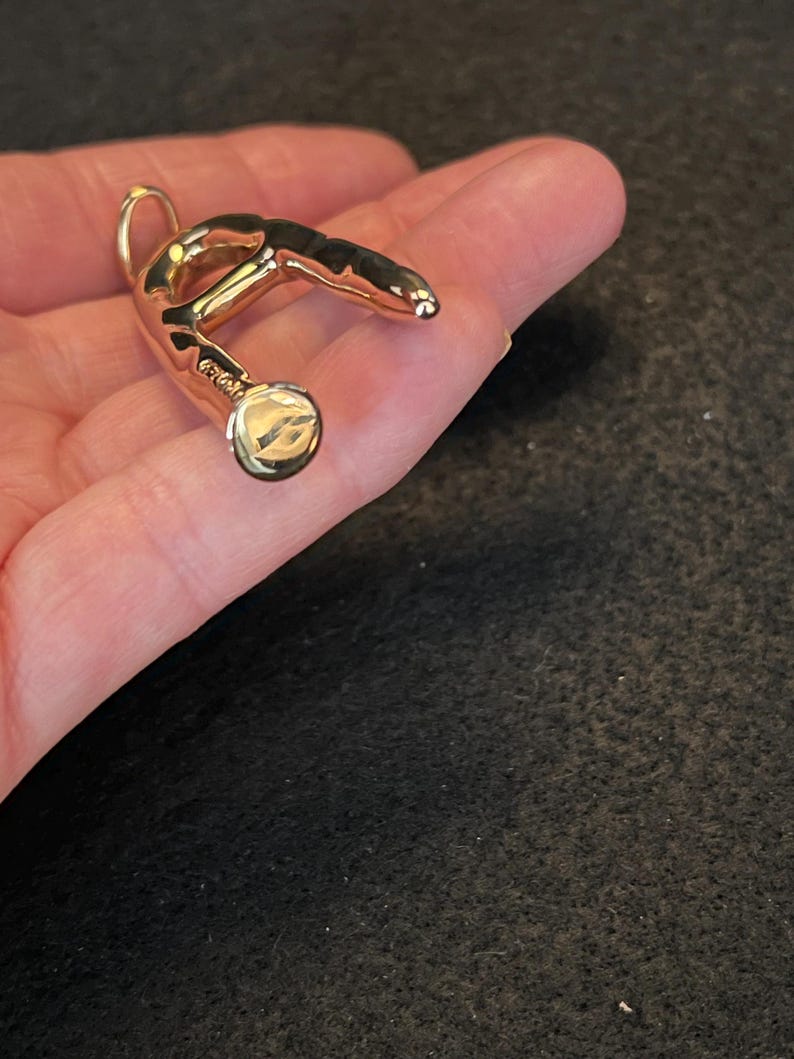 Puede incluir: Un colgante dorado con un dise&ntilde;o curvo y abstracto. El colgante tiene un lazo en la parte superior para una cadena y un detalle circular en la parte inferior. La superficie es lisa y reflectante, con un peque&ntilde;o detalle grabado.