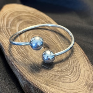 Op de afbeelding: Een zilverkleurige manchetarmband met twee bolvormige accenten. De armband heeft een eenvoudig, open ontwerp met een gladde, gepolijste afwerking. De bollen zijn gelijkmatig verdeeld en geven de sieraden een moderne uitstraling.