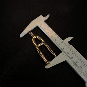 Puede incluir: Un colgante dorado con forma de la letra "A" se mide con un calibrador. El colgante tiene una superficie texturizada y est&aacute; suspendido sobre un fondo oscuro. El calibrador muestra una medida de aproximadamente 3,8 cm.