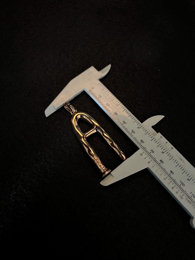 Puede incluir: Un colgante dorado con forma de la letra "A" se mide con un calibrador. El colgante tiene una superficie texturizada y est&aacute; suspendido sobre un fondo oscuro. El calibrador muestra una medida de aproximadamente 3,8 cm.