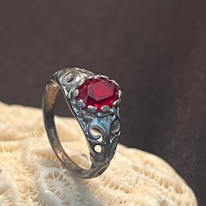 May include: A silver ring featuring an oval-cut deep red gemstone set in a pronged mounting. The band displays an intricate openwork filigree pattern and is stamped with the word STERLING on the inner surface of the metal shank.