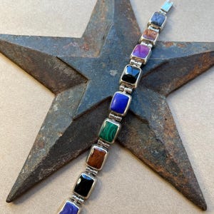 Pode incluir: Uma pulseira de prata com pedras preciosas quadradas multicoloridas. As pedras preciosas incluem azul, verde, preto, castanho, roxo e azul claro. A pulseira é exibida em uma estrela de metal rústico.
