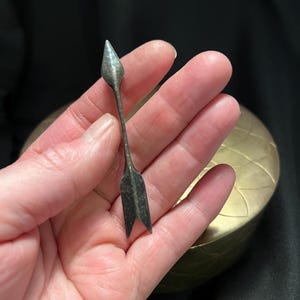May include: A small, antique silver arrow with a pointed tip and split tail feathers. The arrow is held in a hand against a dark background, with a gold-colored, round container visible in the background. The arrow is likely a decorative item or a small piece of jewelry.