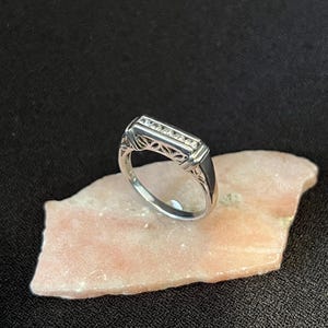 May include: A silver ring with a rectangular top set with small, clear stones. The ring has an ornate, openwork design on the sides and is displayed on a light pink, textured surface against a black background.