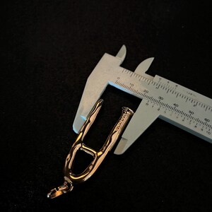 Puede incluir: Un colgante de oro rosa con un dise&ntilde;o abierto &uacute;nico, sostenido por un calibrador plateado. El colgante tiene un lazo en la parte inferior y una superficie texturizada. El calibrador mide en cent&iacute;metros.