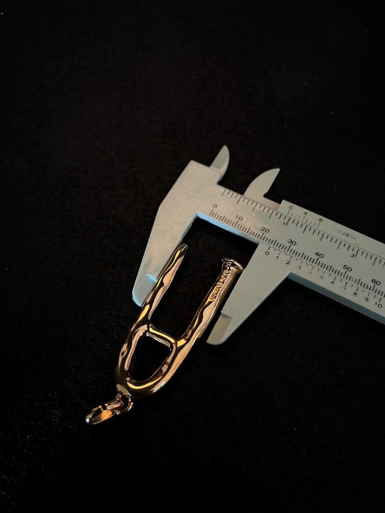 Puede incluir: Un colgante de oro rosa con un dise&ntilde;o abierto &uacute;nico, sostenido por un calibrador plateado. El colgante tiene un lazo en la parte inferior y una superficie texturizada. El calibrador mide en cent&iacute;metros.