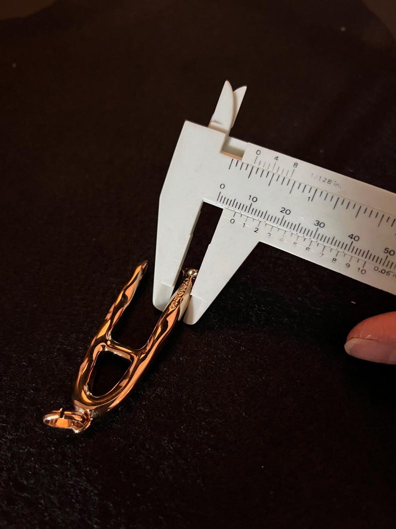 Puede incluir: Un objeto met&aacute;lico de color oro rosa se mide con un calibrador blanco. El objeto tiene un dise&ntilde;o abierto &uacute;nico con brazos curvos y una peque&ntilde;a base redondeada. El calibrador muestra medidas en cent&iacute;metros.