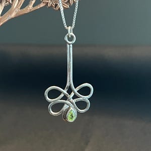 May include: A silver pendant necklace featuring a floral design with a pear-shaped, light green gemstone at the center. The pendant hangs from a delicate silver chain. The necklace is displayed against a blurred background.