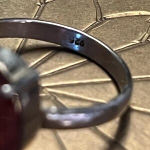 Pode incluir: Close-up de um anel de prata com uma grande pedra preciosa vermelha oval. A banda do anel est&aacute; marcada com "925". O anel est&aacute; sobre uma superf&iacute;cie texturizada castanho-dourada.