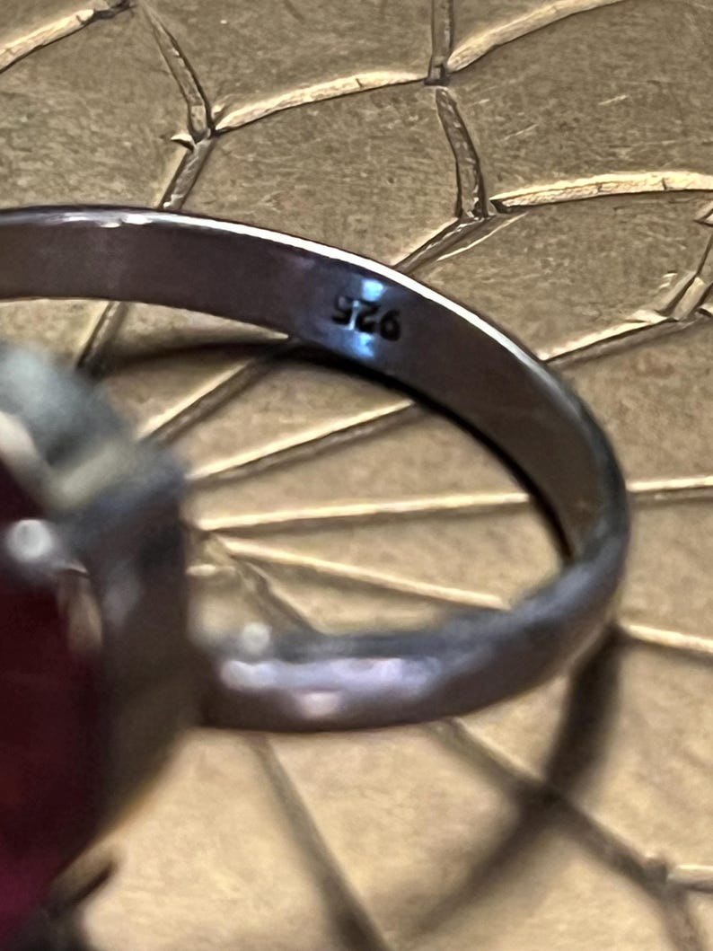 Pode incluir: Close-up de um anel de prata com uma grande pedra preciosa vermelha oval. A banda do anel est&aacute; marcada com "925". O anel est&aacute; sobre uma superf&iacute;cie texturizada castanho-dourada.