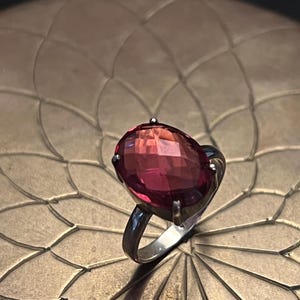 Pode incluir: Um anel de prata com uma grande pedra preciosa rosa-avermelhada de corte oval e facetado. O anel est&aacute; sobre uma superf&iacute;cie texturizada castanho-dourada, criando um contraste marcante. A pedra preciosa &eacute; o ponto focal.