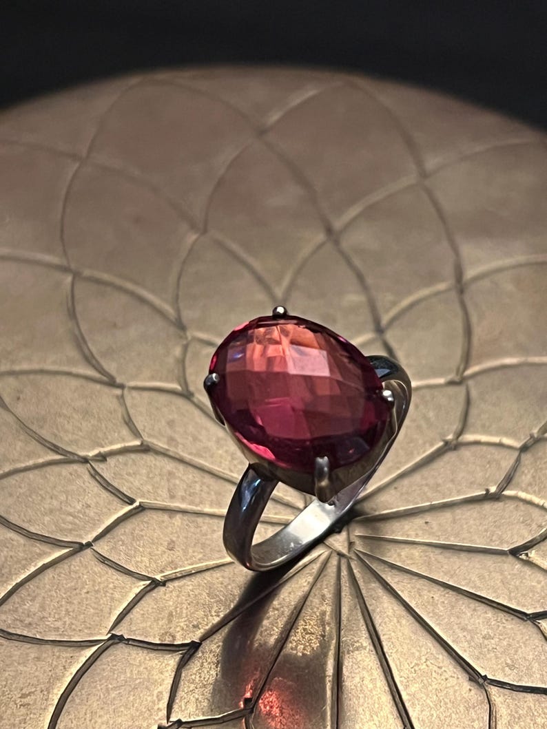 Pode incluir: Um anel de prata com uma grande pedra preciosa rosa-avermelhada de corte oval e facetado. O anel est&aacute; sobre uma superf&iacute;cie texturizada castanho-dourada, criando um contraste marcante. A pedra preciosa &eacute; o ponto focal.