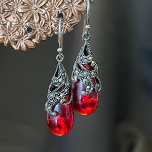 May include: A pair of drop earrings featuring teardrop-shaped translucent red stones suspended from silver-toned metal settings. The metalwork displays intricate filigree patterns and small, dark, faceted stone accents. The earrings hang from silver hooks.