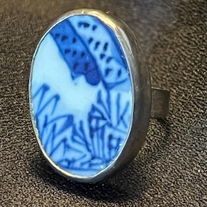 Puede incluir: Anillo ovalado con banda de plata. El anillo presenta una incrustación de cerámica azul claro y blanca con un diseño floral azul. El diseño del anillo recuerda a la porcelana antigua.