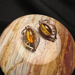 May include: A pair of vintage earrings featuring oval amber-colored stones set in ornate silver-toned metal. The earrings have a leaf-like design with decorative details. The jewelry is displayed on a marbled, multi-colored surface.