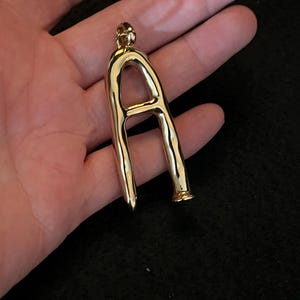 Puede incluir: Un colgante dorado con forma de la letra "A". El colgante tiene una superficie lisa y pulida y un peque&ntilde;o lazo en la parte superior para una cadena. La parte inferior del colgante tiene una punta y una base acampanada.
