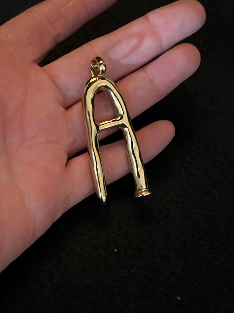 Puede incluir: Un colgante dorado con forma de la letra "A". El colgante tiene una superficie lisa y pulida y un peque&ntilde;o lazo en la parte superior para una cadena. La parte inferior del colgante tiene una punta y una base acampanada.
