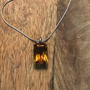 May include: A necklace featuring a rectangular, faceted amber-colored gemstone pendant set in a silver-toned metal frame, suspended from a thin, flexible silver-toned snake chain that curves gracefully across the frame of the image.