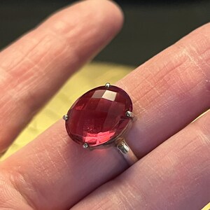 Pode incluir: Um anel de prata com uma grande pedra preciosa vermelha escura, facetada e de forma oval. A pedra &eacute; mantida no lugar por quatro pequenas garras. A banda do anel &eacute; de prata polida.