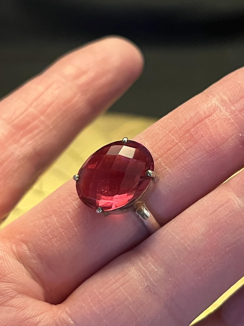 Pode incluir: Um anel de prata com uma grande pedra preciosa vermelha escura, facetada e de forma oval. A pedra &eacute; mantida no lugar por quatro pequenas garras. A banda do anel &eacute; de prata polida.