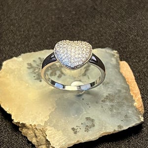 May include: A silver ring with a heart-shaped design. The heart is covered in small, sparkling stones. The ring is set on a light-colored, textured surface against a dark background. The ring's band is smooth and shiny.