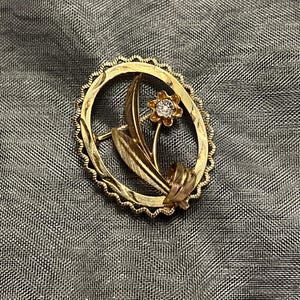 May include: A gold-toned oval brooch featuring a floral design. The brooch has a textured frame with a flower and leaf motif. The flower has a central diamond-like stone.