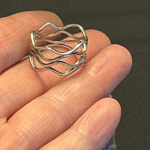 Puede incluir: Un anillo de color plateado con un diseño de múltiples capas compuesto por varias bandas finas y onduladas interconectadas. El metal tiene un acabado liso y pulido, creando una estructura calada continua alrededor del dedo.