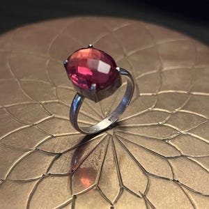 May include: A silver ring with a large, faceted, oval-shaped gemstone. The gemstone transitions from a deep pink to a lighter red hue. The ring is set on a decorative, gold-colored surface.
