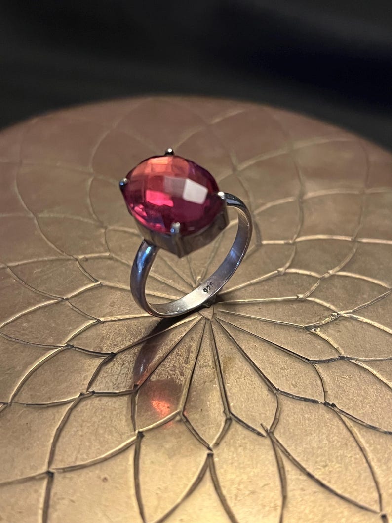 Pode incluir: Um anel de prata com uma grande pedra preciosa oval e facetada. A pedra preciosa transita de um rosa profundo para um tom vermelho mais claro. O anel est&aacute; sobre uma superf&iacute;cie decorativa dourada.