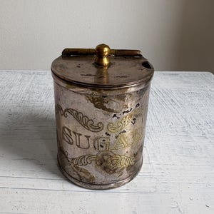 May include: A cylindrical metal sugar canister with a tarnished silver-tone finish and a hinged lid featuring a brass-coloured knob. The word SUGAR is embossed on the front, framed by decorative scrollwork patterns in a brass-coloured finish.