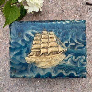 May include: A rectangular bar of soap featuring a raised, cream-coloured relief of a sailing ship with multiple masts and sails. The soap has a marbled, swirling pattern of deep blue, teal, and white, creating the appearance of ocean waves.