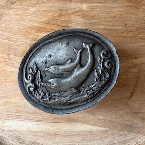 May include: An oval-shaped metal belt buckle featuring a raised relief design of two dolphins swimming among seaweed and bubbles. The surface has a dark, weathered grey finish with a textured background that highlights the aquatic scene.