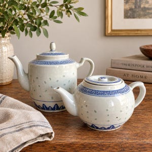 May include: Two ceramic teapots with a light gray glaze, featuring blue hand-painted geometric patterns around the rims and bases, and small blue dots scattered across the bodies. Both teapots have matching lids with small knobs and curved handles.