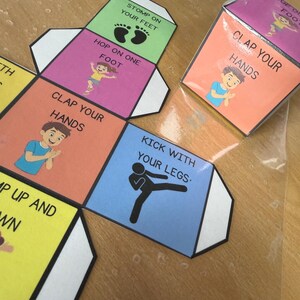 Puede incluir: Dados de actividad coloridos con instrucciones como "Aplaudir" y "Patear con las piernas". Los dados están hechos de papel plegado con ilustraciones de niños realizando las acciones.