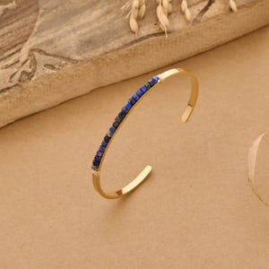 May include: A gold-colored cuff bracelet with a row of small, dark blue and black beads. The bracelet has a simple, open design and is displayed on a neutral-toned surface. The beads are evenly spaced along the top of the bracelet.