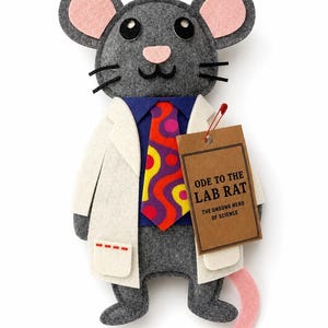 May include: A gray felt rat plush toy wearing a white lab coat over a blue shirt and a colorful patterned tie. The rat has pink inner ears, a pink tail, and black eyes. A brown paper tag attached with a red pin reads: ODE TO THE LAB RAT THE UNSUNG HERO OF SCIENCE.